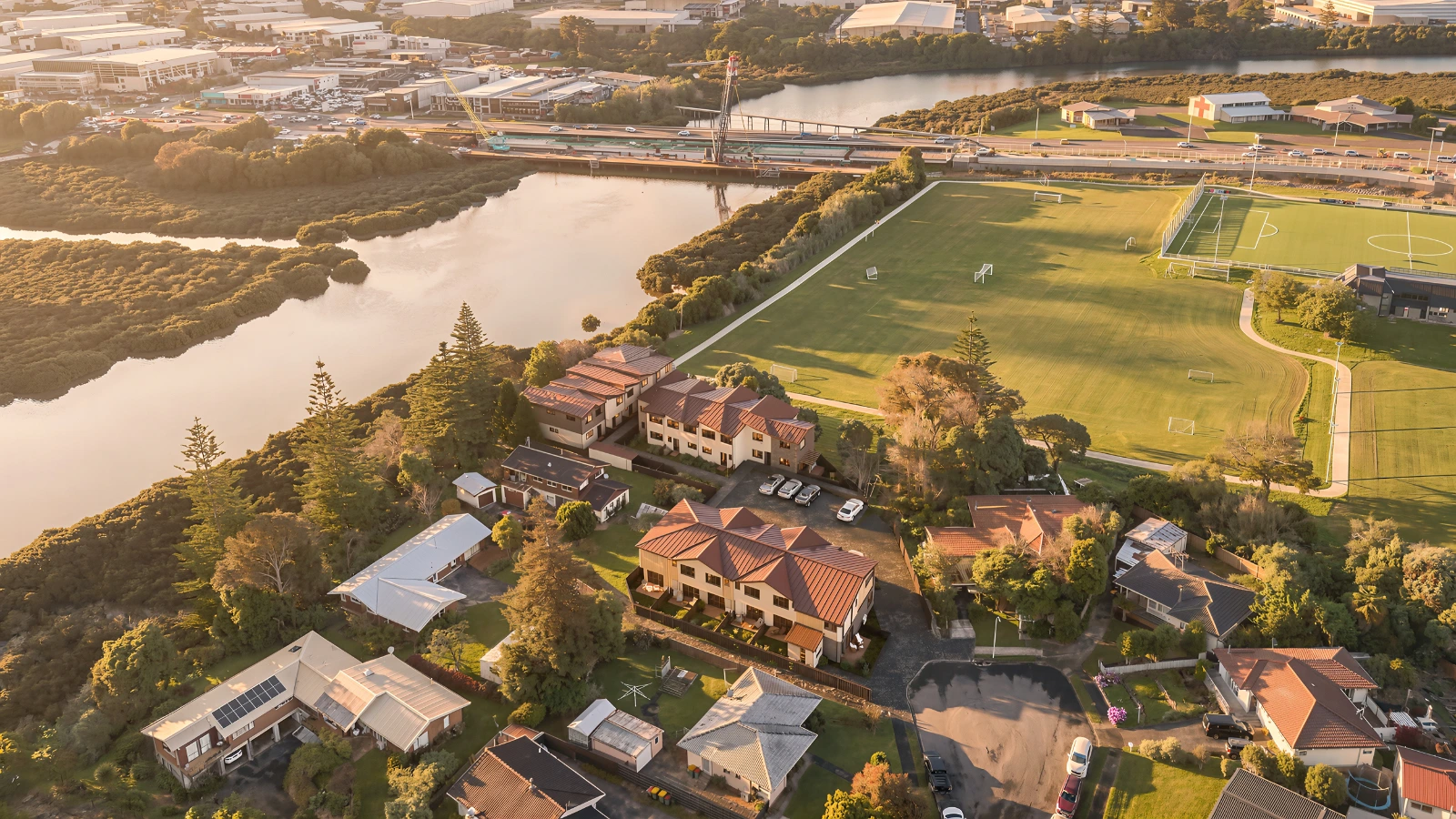 High-altitude aerial photomontage showcasing a new residential townhouse development integrated into a real-world landscape at sunset . The 3D architectural visualization features multiple multi-story residential buildings with distinct brown pitched roofs nestled near a winding river and expansive green sports fields[cite: 26]. The background reveals a sprawling industrial park, a bridge crossing the water, and distant rolling hills under a warm golden hour sky.