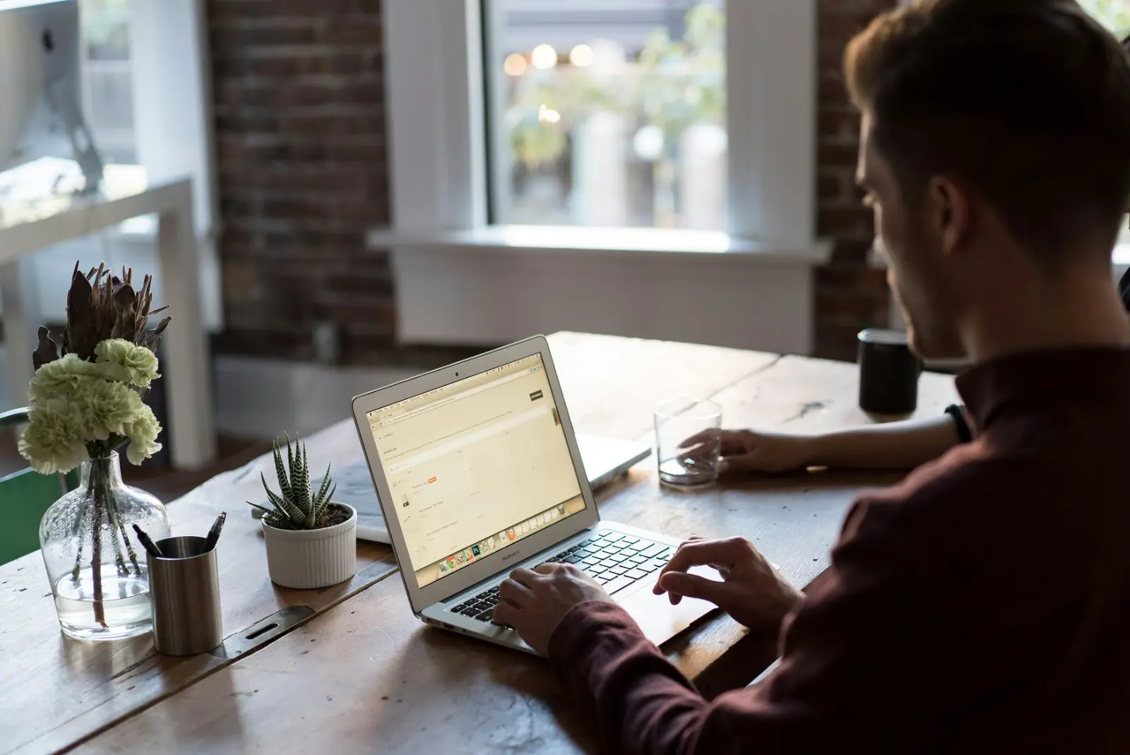 High-quality lifestyle photograph featuring a creative professional typing on a silver laptop at a rustic wooden desk in a bright, modern office space. The scene includes a glass of water and decorative plants, illustrating the focused, day-to-day workflow and environment at Our-Inspiration.