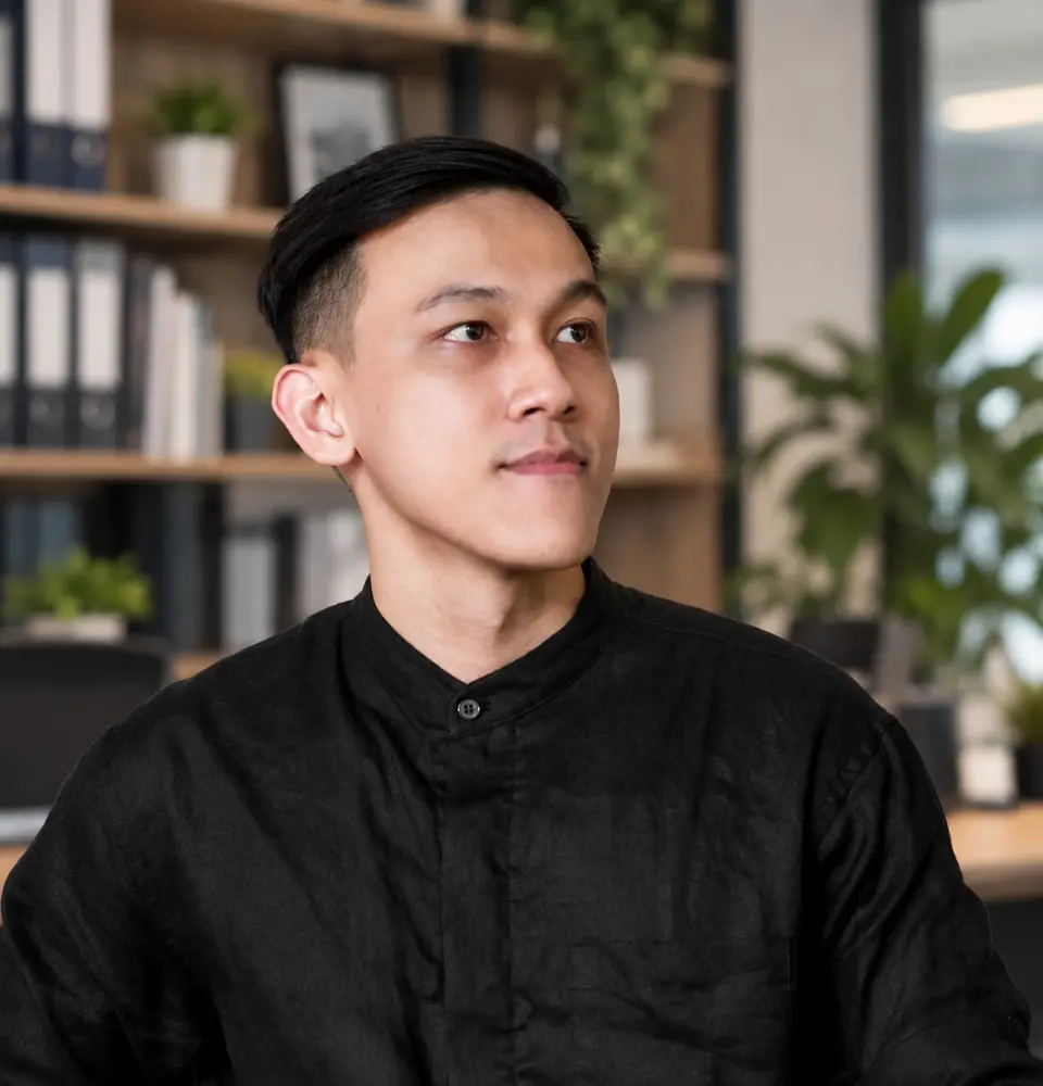 Professional portrait photograph of Huy Nguyen, Exterior Leader of Our-Inspiration, wearing a black shirt, posing in a modern office environment with a bookshelf background.