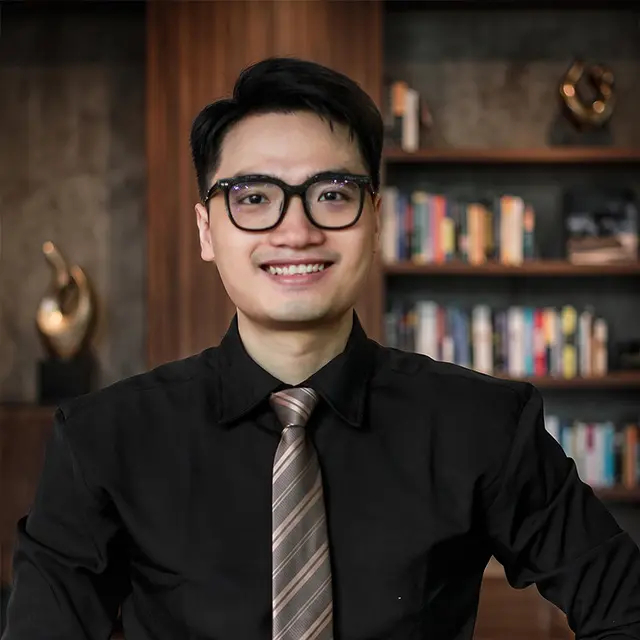 Professional portrait photograph of Kha, CEO and founder of Our-Inspiration, wearing glasses, a black shirt, and a tie, posing with a warm executive office background featuring a wooden bookshelf.