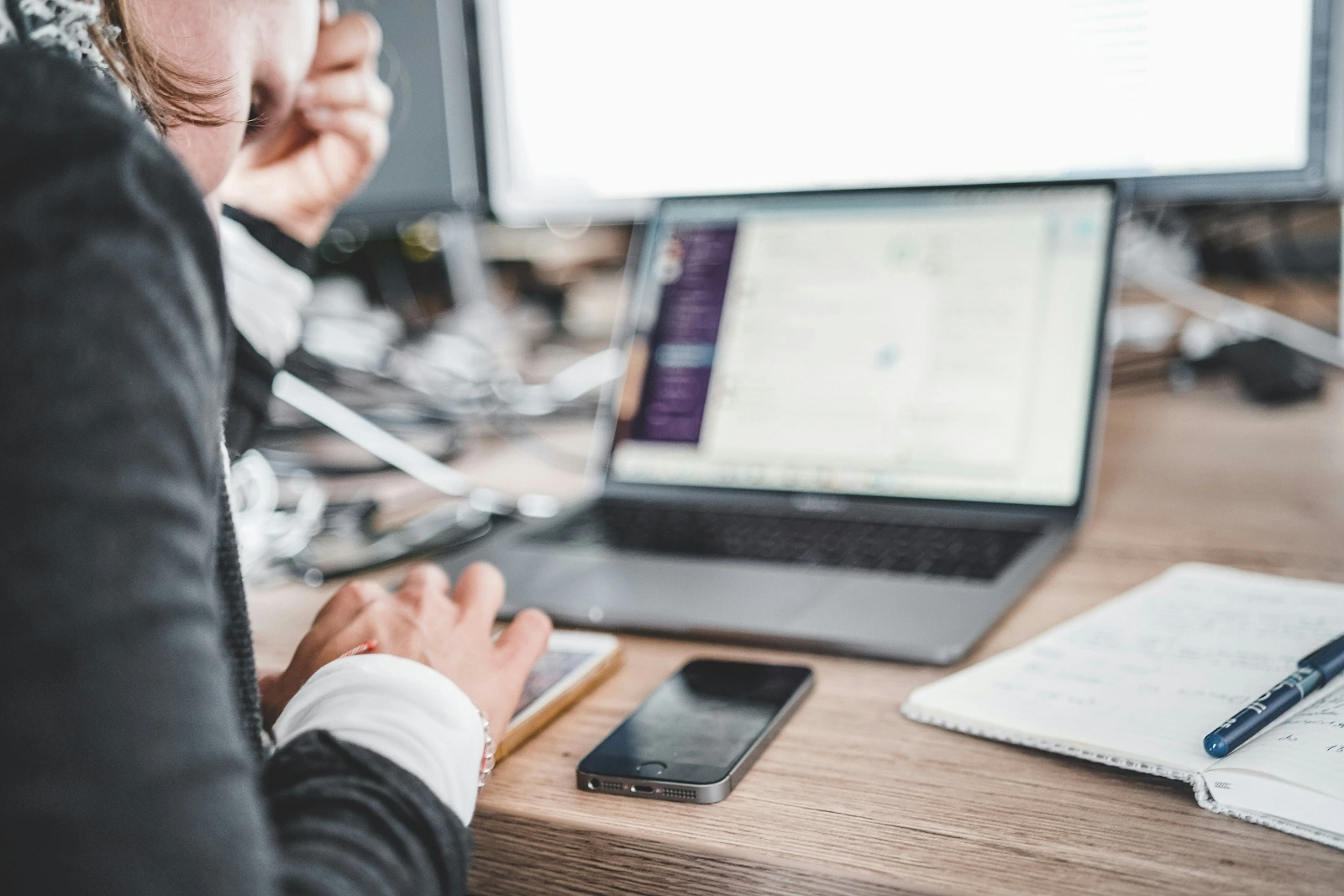 High-quality lifestyle photography capturing a professional working at a wooden office desk from an over-the-shoulder perspective. The active workspace includes an open laptop, a black smartphone, an open notebook, and a blue pen. The shallow depth of field softly blurs the laptop screen and background monitor, emphasizing a modern, focused business environment.
