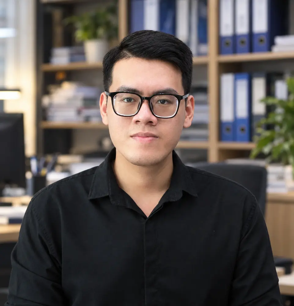 Professional portrait photograph of Truong Bui, Secretary of Our-Inspiration, wearing glasses and a black shirt, posing in an office environment with file binders on a bookshelf in the background.